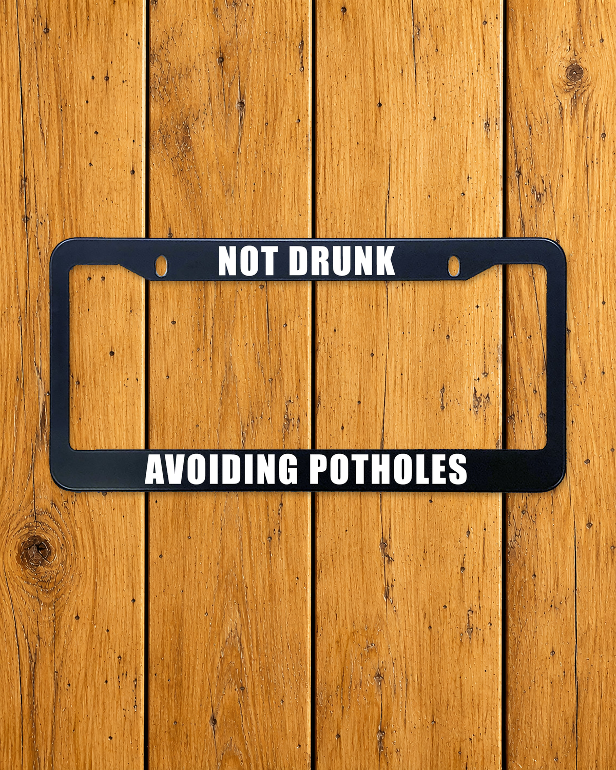 Not Drunk, Avoiding Potholes license plate frame shown on a warm wood background