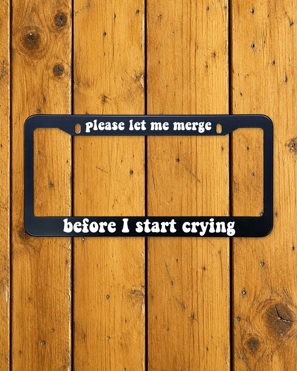 Please Let Me Merge, Before I Start Crying license plate frame shown on a warm wood background